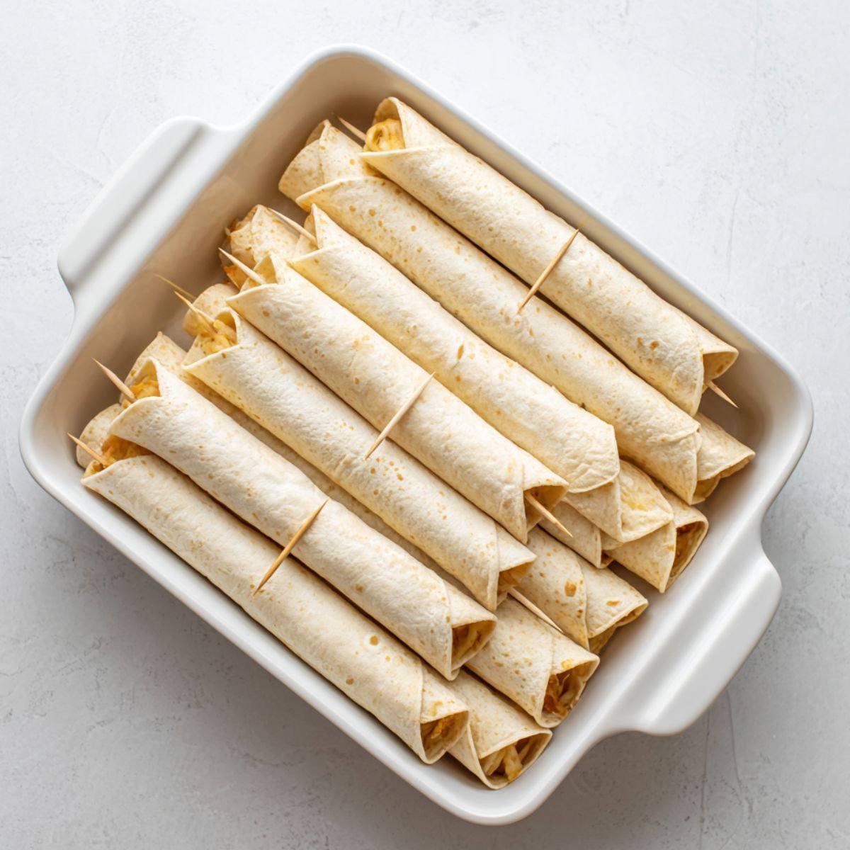 Uncooked homemade chicken flautas neatly rolled and secured with toothpicks, arranged in rows inside a white ceramic baking dish on a light kitchen counter.