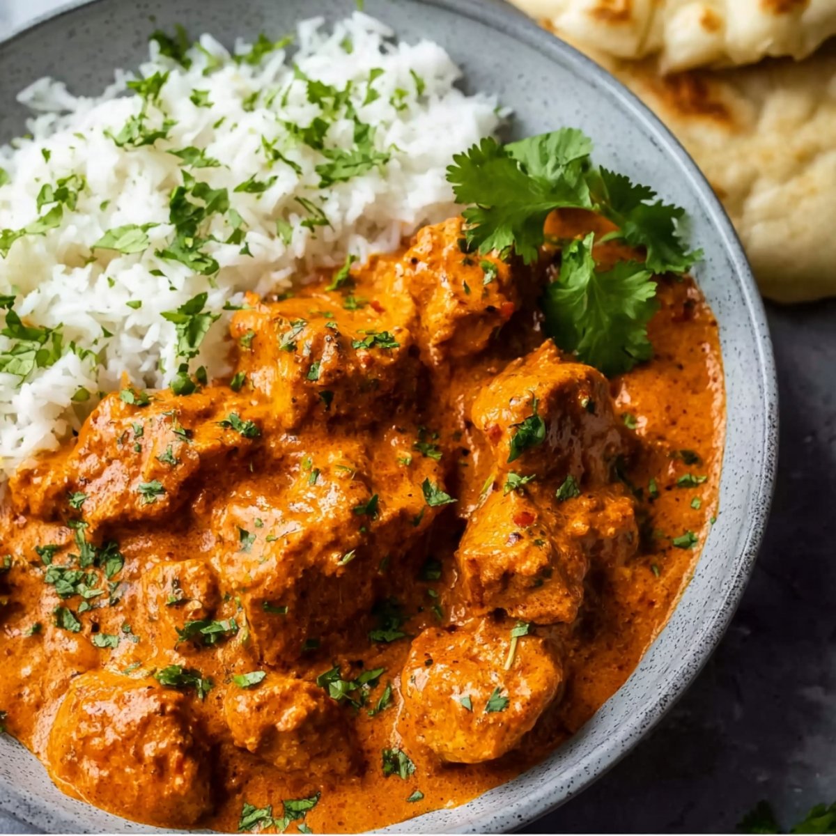 A bowl of homemade butter chicken served with basmati rice and fresh cilantro, with naan on the side.