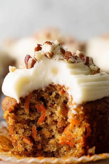 A Carrot Cake Cupcakes with cream cheese frosting and pecans, revealing its moist, tender interior filled with grated carrots.