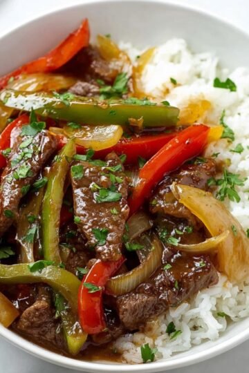 Pepper steak with colorful bell peppers, onions, and tender beef slices, served over white rice and garnished with chopped parsley.