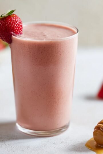 Strawberry smoothie in a glass with a fresh strawberry garnish and honey nearby.