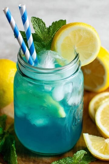A mason jar filled with Blue Raspberry Lemonade, garnished with mint leaves and lemon slices, placed on a wooden plate.