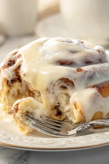 Close-up of a warm Cinnabon Cinnamon Rolls drenched in creamy icing with a fork cutting into the soft, fluffy layers, icing melting and pooling on the plate