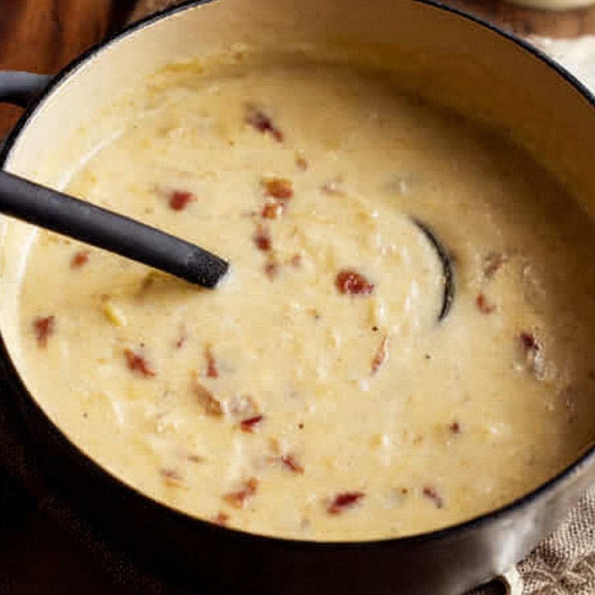 A creamy potato soup with bacon bits in a pot, stirred with a black spoon.