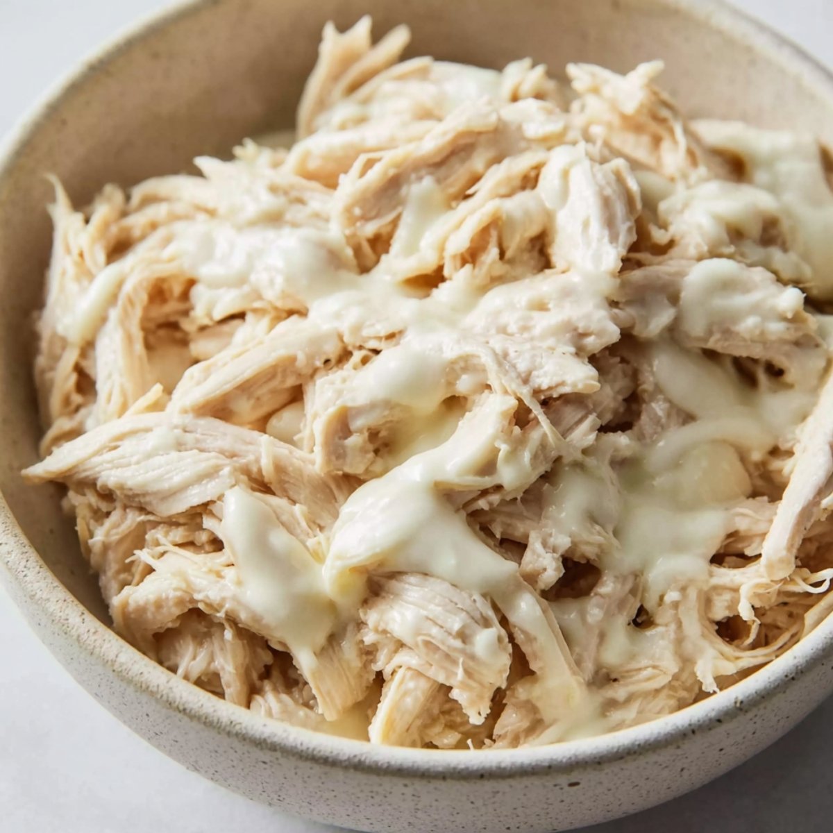 Shredded cooked chicken in a bowl with creamy sauce drizzled on top.