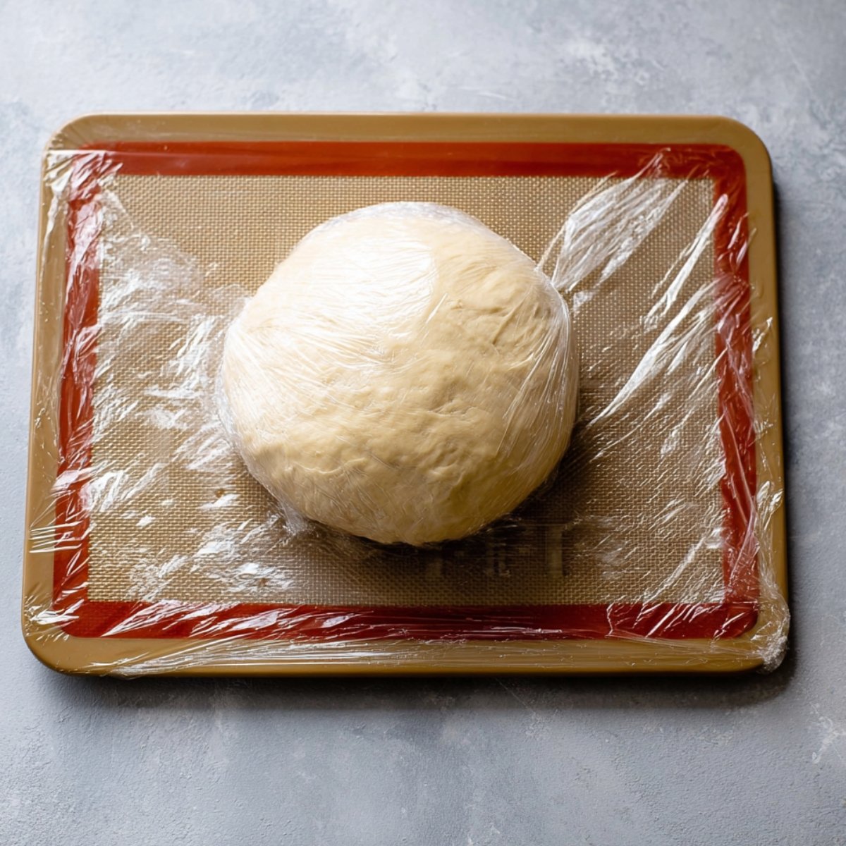 A ball of croissant dough, wrapped in plastic, resting on a baking mat.