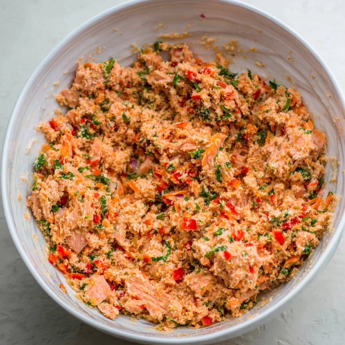 A bowl filled with a mixture of salmon, breadcrumbs, red peppers, and herbs, all thoroughly combined to create the base for homemade salmon patties.