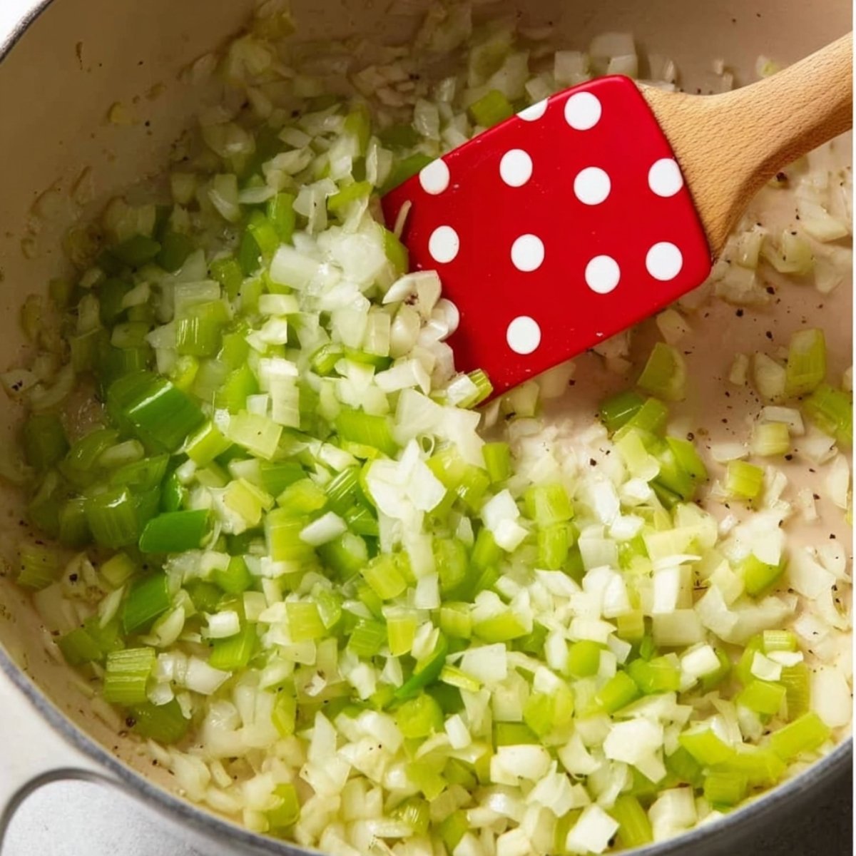 Chopped onions and celery sautéing in a pot with a red spatula.